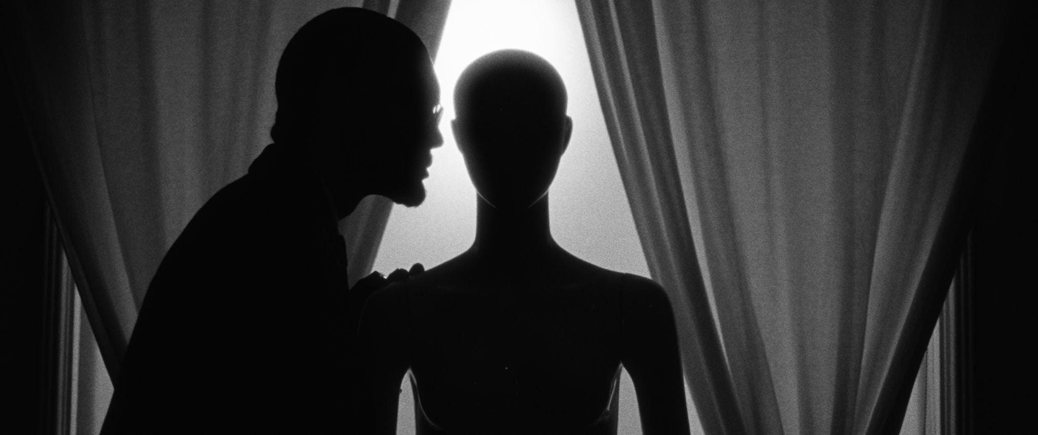 A man in silhouette whispers to a featureless mannequin before glowing curtains.
